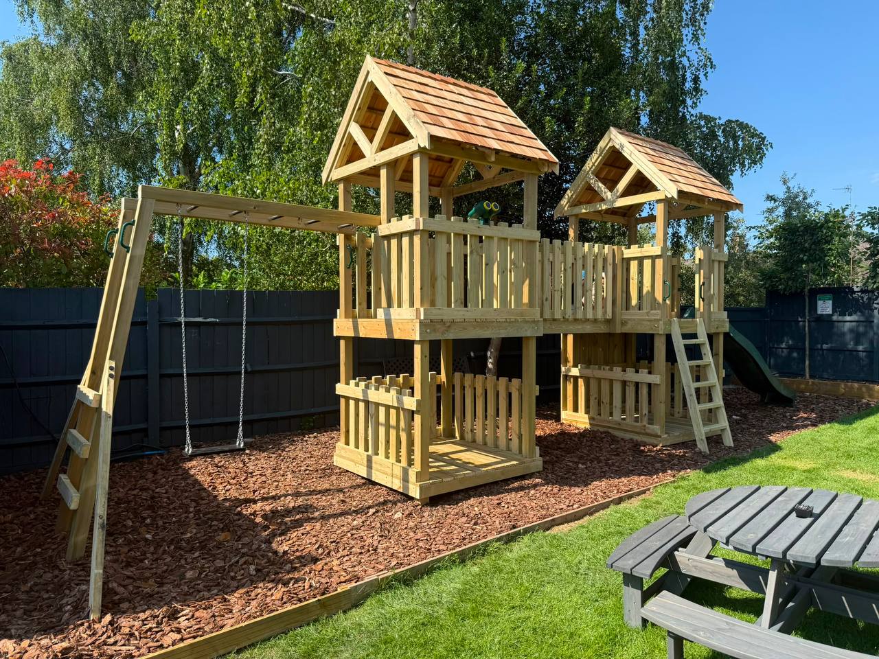 Outdoor play area at The Side Quest with wooden climbing towers, swings, slide and lawn seating.